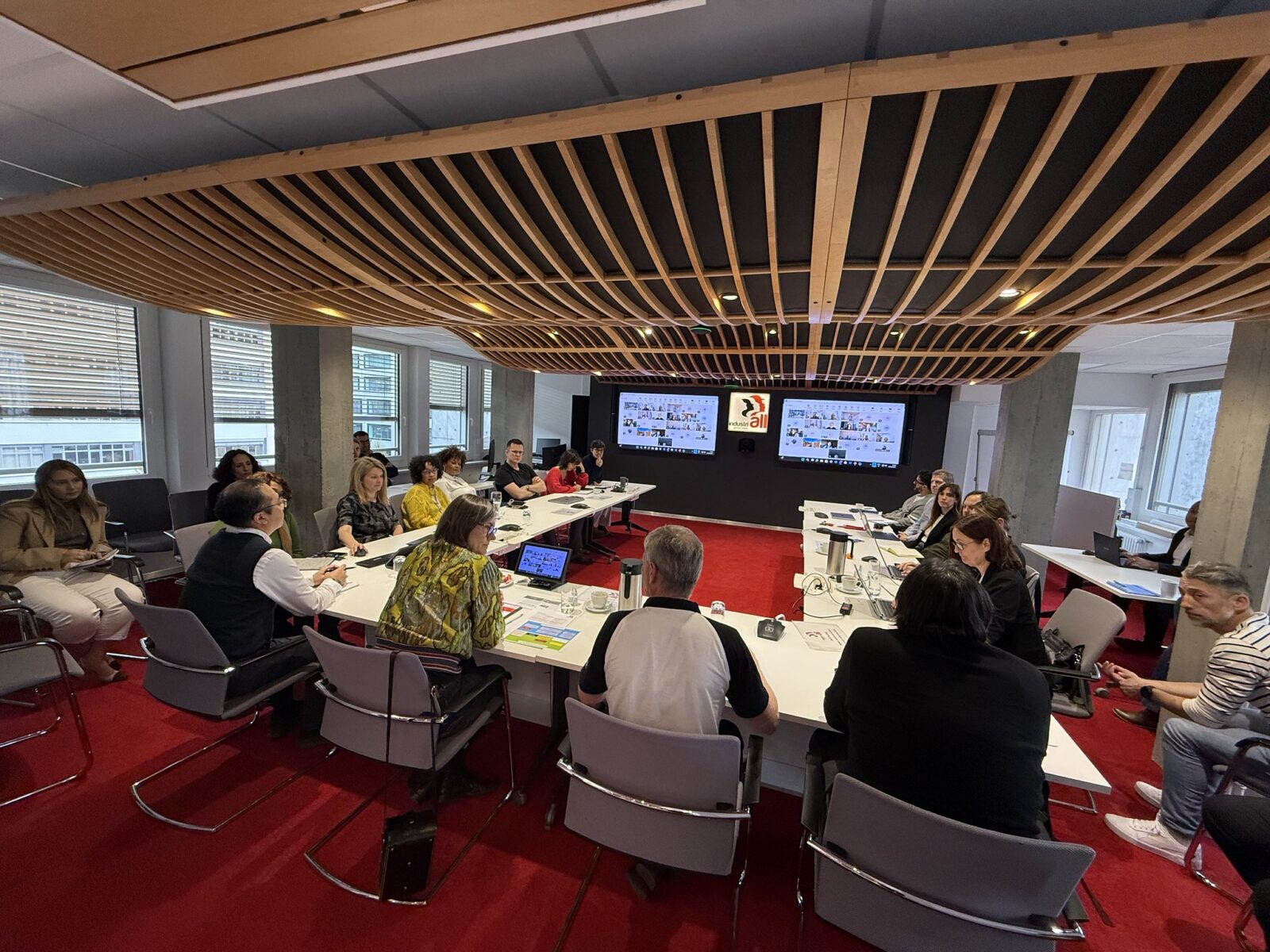 Wide-angle view of a meeting at IndustriALL Global Union headquarters in Geneva, with staff and guests seated around a large table, IndustriALL logo visible on screens, 25 March 2026